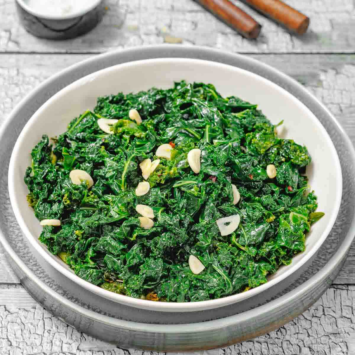 A white bowl filled with sautéed kale and sliced garlic, placed on a plate on a rustic wooden surface.