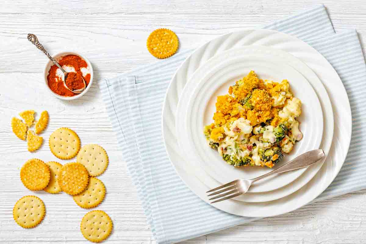 A serving of broccoli casserole with a crispy topping and melted cheese on a white plate with a fork, placed on stacked plates over a striped napkin.