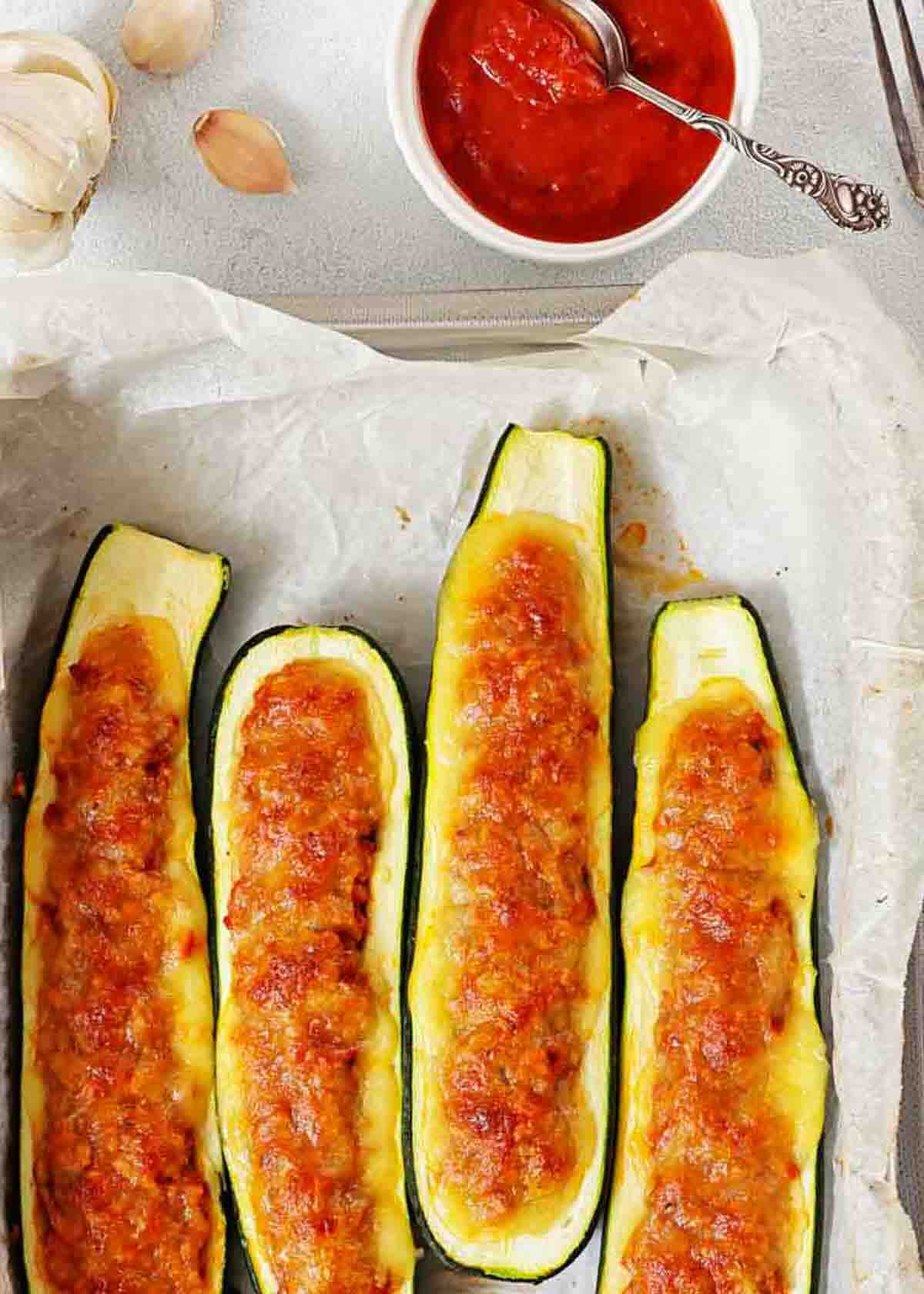 Four baked zucchini boats filled with a golden, cheesy tomato mixture on parchment paper, with a bowl of tomato sauce and a spoon in the background.