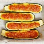 Four zucchini halves stuffed with a seasoned filling and topped with melted cheese, arranged in a baking dish lined with parchment paper.