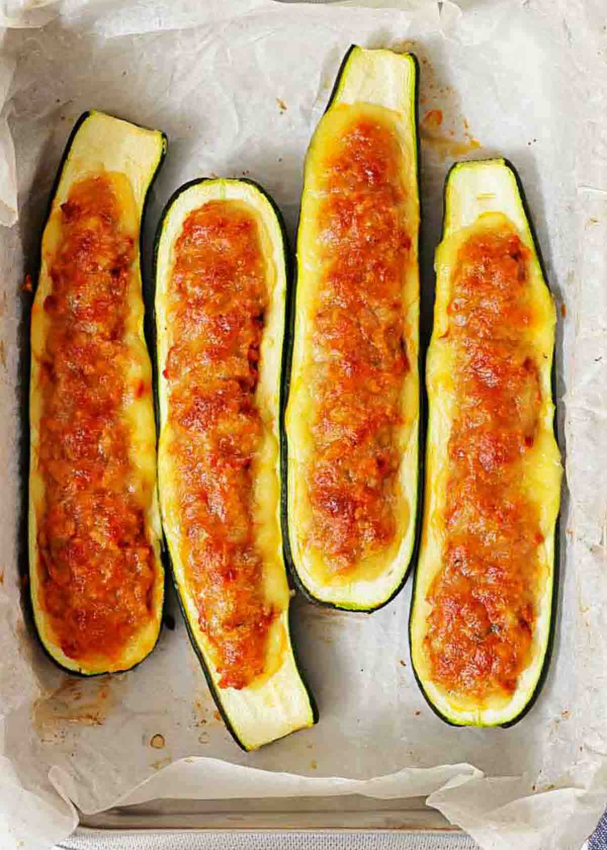 Four baked zucchini boats filled with a browned, cheesy stuffing are arranged on parchment paper in a baking dish.