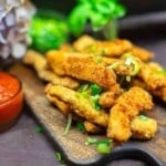A pile of breaded and fried chicken strips garnished with chopped green onions on a wooden board, with a small bowl of red dipping sauce on the side.