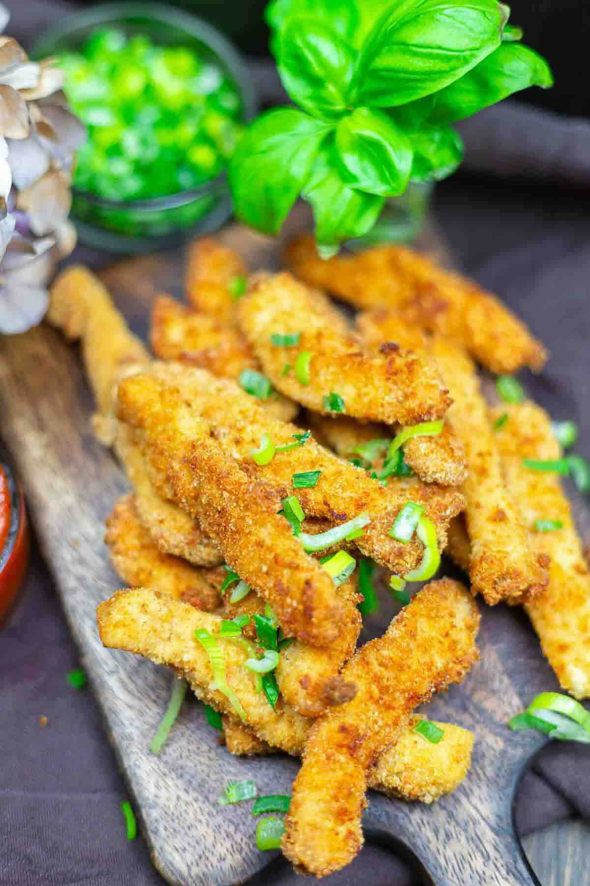 Breaded and fried mushroom strips garnished with chopped green onions, served on a wooden board with fresh basil and a bowl of green sauce in the background.