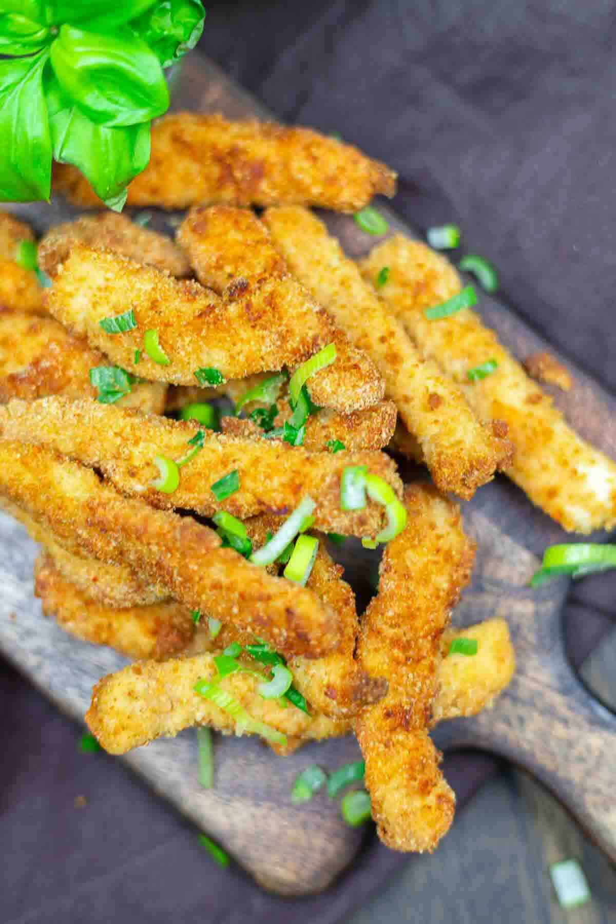 A pile of breaded and fried zucchini sticks garnished with chopped green onions on a wooden serving board.