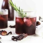 A glass of dark red iced drink with ice cubes, garnished with a sprig of rosemary, surrounded by dried hibiscus petals.