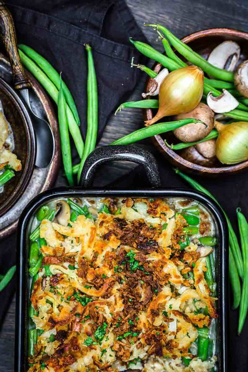 A baked green bean casserole topped with crispy fried onions is surrounded by fresh green beans, mushrooms, and onions on a dark surface.