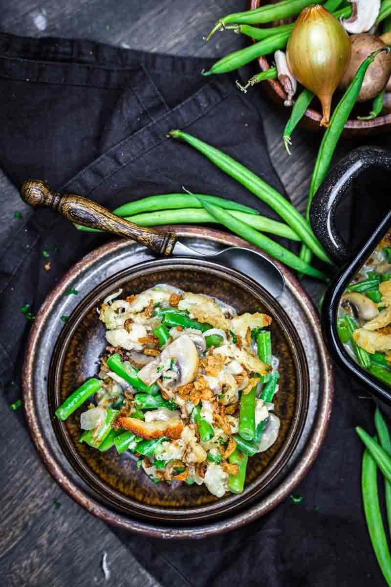 A bowl of green bean casserole with mushrooms and fried onions, surrounded by fresh green beans and an onion on a dark cloth.