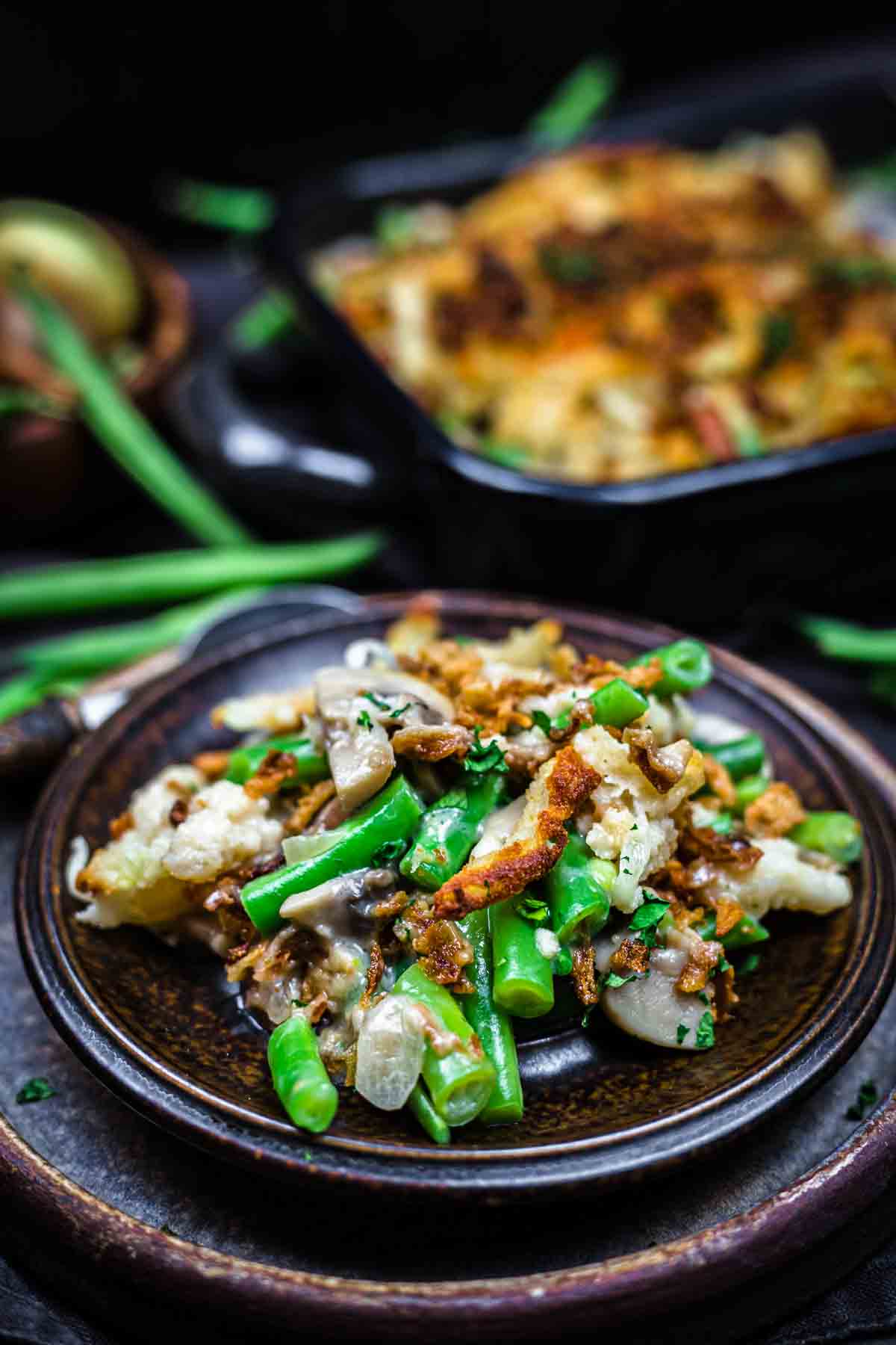 A plate of green bean casserole topped with crispy fried onions, featuring green beans, mushrooms, and a creamy sauce. A serving dish is visible in the background.
