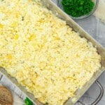A metal tray filled with scrambled eggs, surrounded by a bowl of chopped herbs and a slice of bread on a wooden surface.