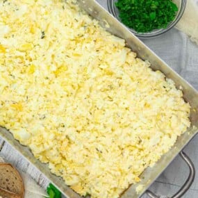 A metal tray filled with scrambled eggs, surrounded by a bowl of chopped herbs and a slice of bread on a wooden surface.