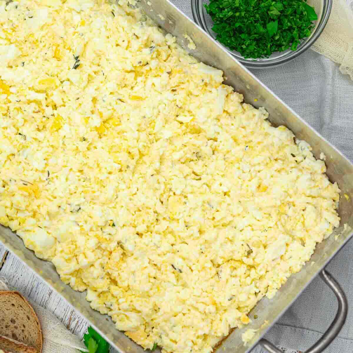 A metal tray filled with scrambled eggs, surrounded by a bowl of chopped herbs and a slice of bread on a wooden surface.