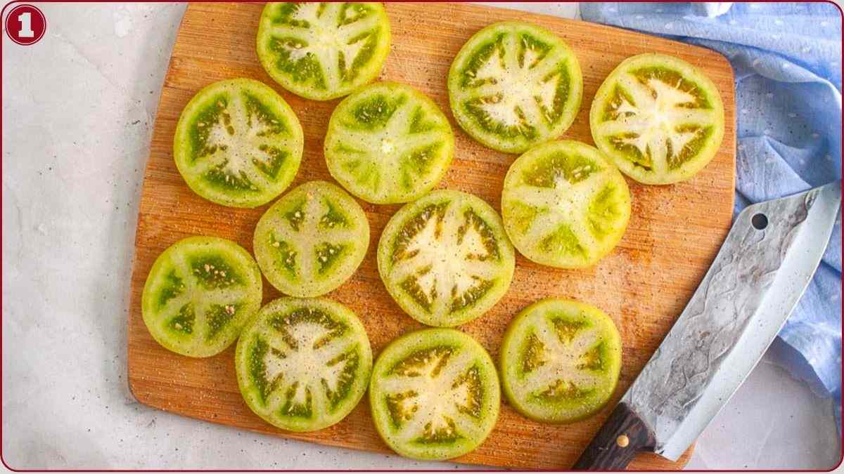 Sliced green tomatoes are arranged on a wooden cutting board next to a large knife, with a blue cloth nearby.
