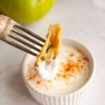 A fork holds a fried slice dipped in a creamy white sauce, above a small ramekin with sauce sprinkled with paprika.
