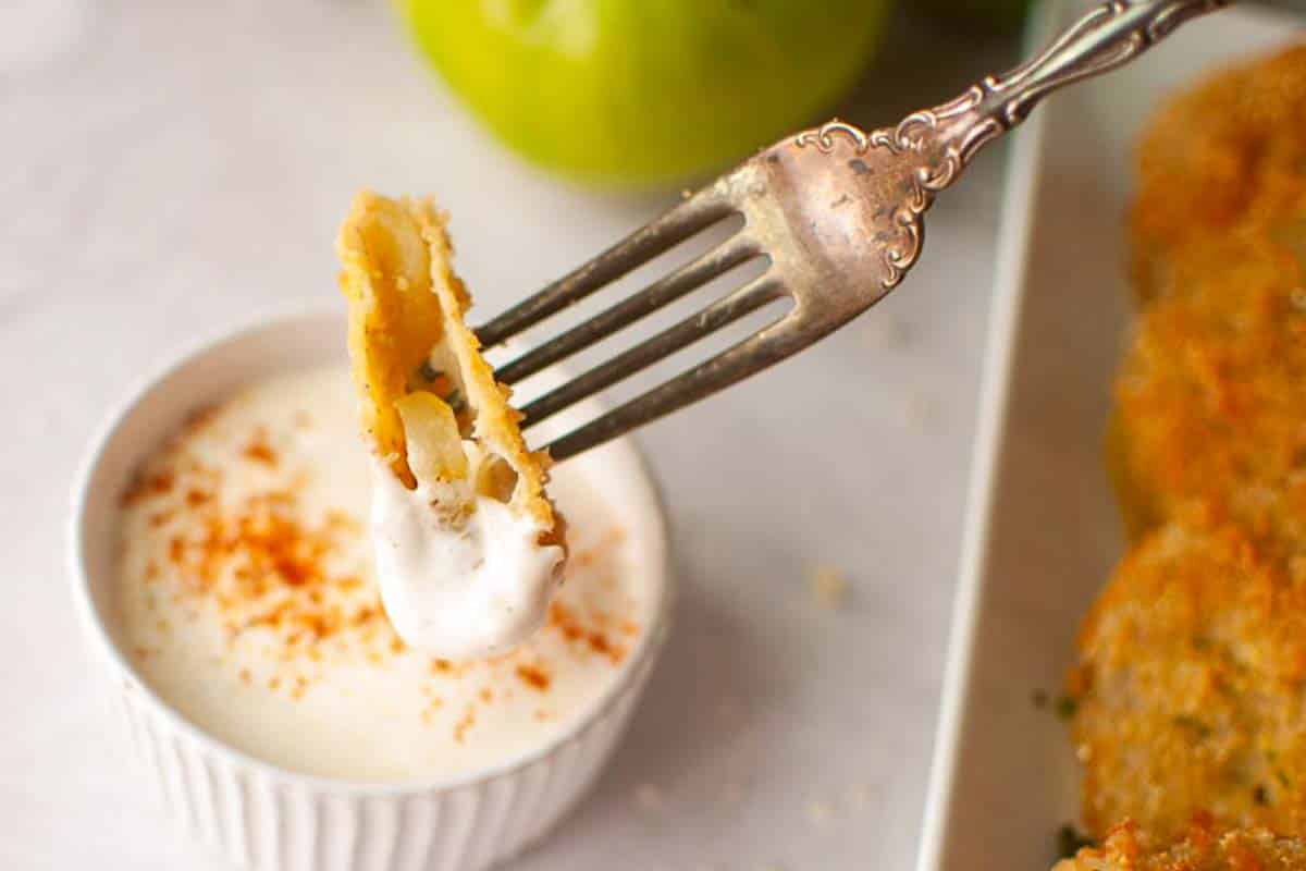 A fork holds a piece of fried green tomato being dipped into a small bowl of creamy white sauce with a sprinkle of paprika.