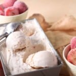 A metal container with scoops of strawberry ice cream and an ice cream scooper, with bowls of frozen strawberries in the background.