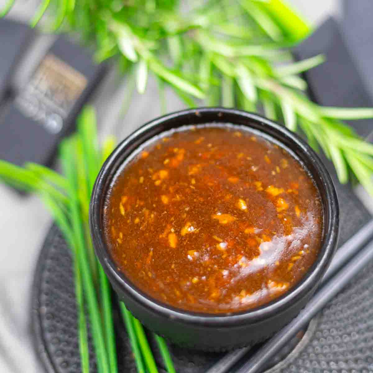 A small black bowl filled with a thick, brown, textured sauce sits on a round black mat, surrounded by green herbs and chopsticks.