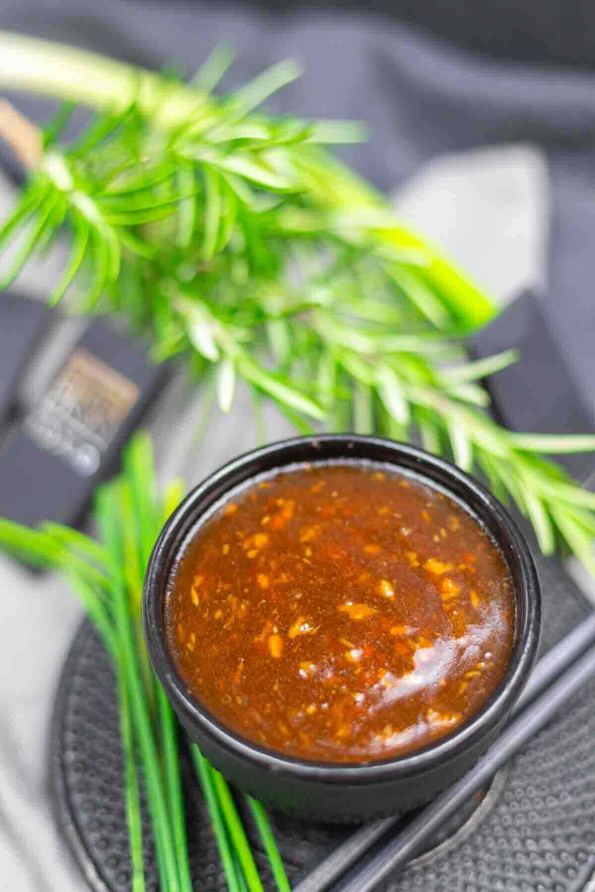 A small black bowl filled with a dark, chunky sauce, surrounded by fresh green herbs on a textured surface.