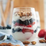 A clear glass filled with layers of whipped cream, blueberries, sliced strawberries, and chopped nuts, placed on a white surface with almonds scattered around.