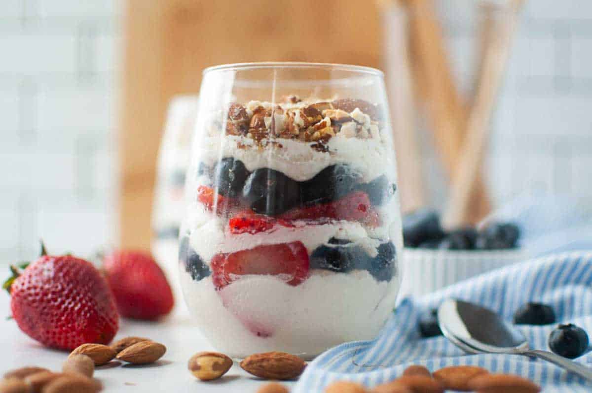 A glass filled with layers of yogurt, blueberries, strawberries, and granola, surrounded by almonds, strawberries, and a spoon on a table.