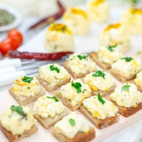 Small pieces of bread topped with an egg salad mixture and garnished with chopped herbs, arranged neatly on a light-colored surface.