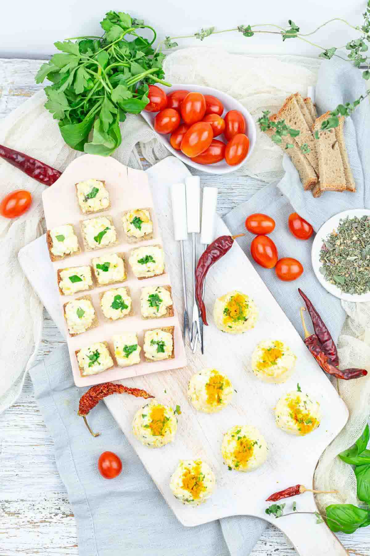 An overhead view of assorted appetizers on a wooden board, surrounded by cherry tomatoes, herbs, bread slices, dried chili peppers, and cutlery.