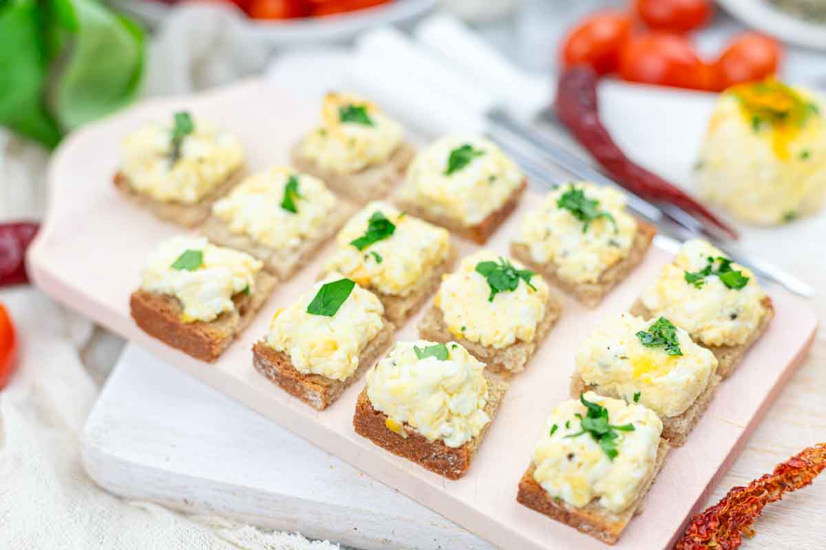 Twelve pieces of bread topped with a creamy egg salad and garnished with herbs are arranged on a white cutting board.