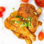 Two pieces of seasoned baked chicken garnished with chopped green onions, served on a white plate with cherry tomatoes and a small dish of sauce in the background.