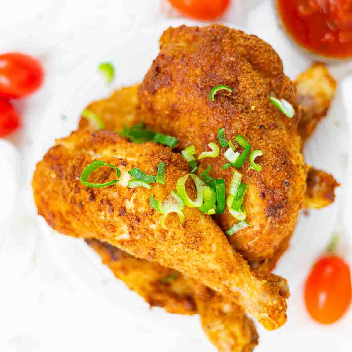 Two pieces of seasoned baked chicken garnished with chopped green onions, served on a white plate with cherry tomatoes and a small dish of sauce in the background.
