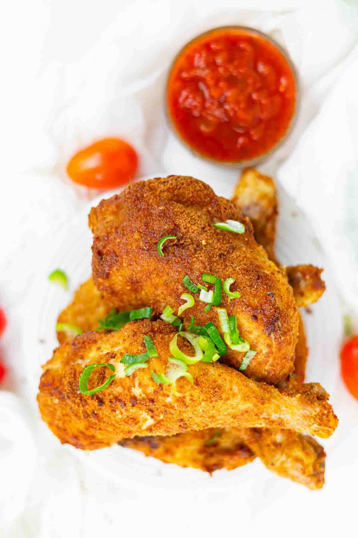 Three seasoned fried chicken legs garnished with chopped green onions, served on a white plate, with a small bowl of red dipping sauce and cherry tomatoes nearby.