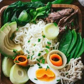 A bowl of ramen noodles topped with avocado, bok choy, shredded beef, snow peas, mushrooms, leeks, carrot ribbons, a soft-boiled egg, and chopped chives.