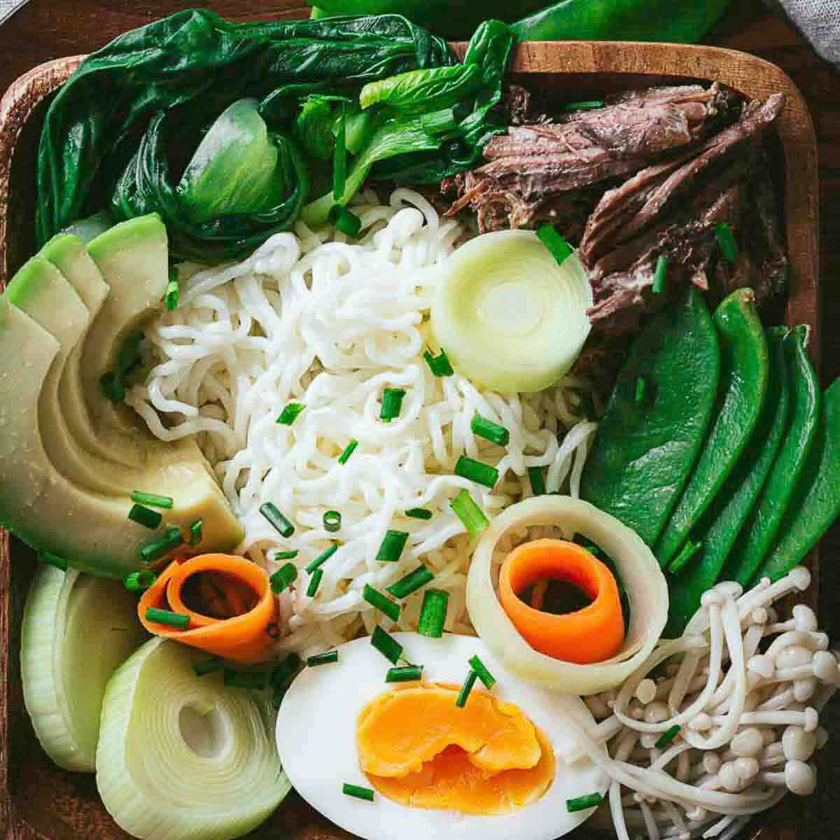A bowl of ramen noodles topped with avocado, bok choy, shredded beef, snow peas, mushrooms, leeks, carrot ribbons, a soft-boiled egg, and chopped chives.