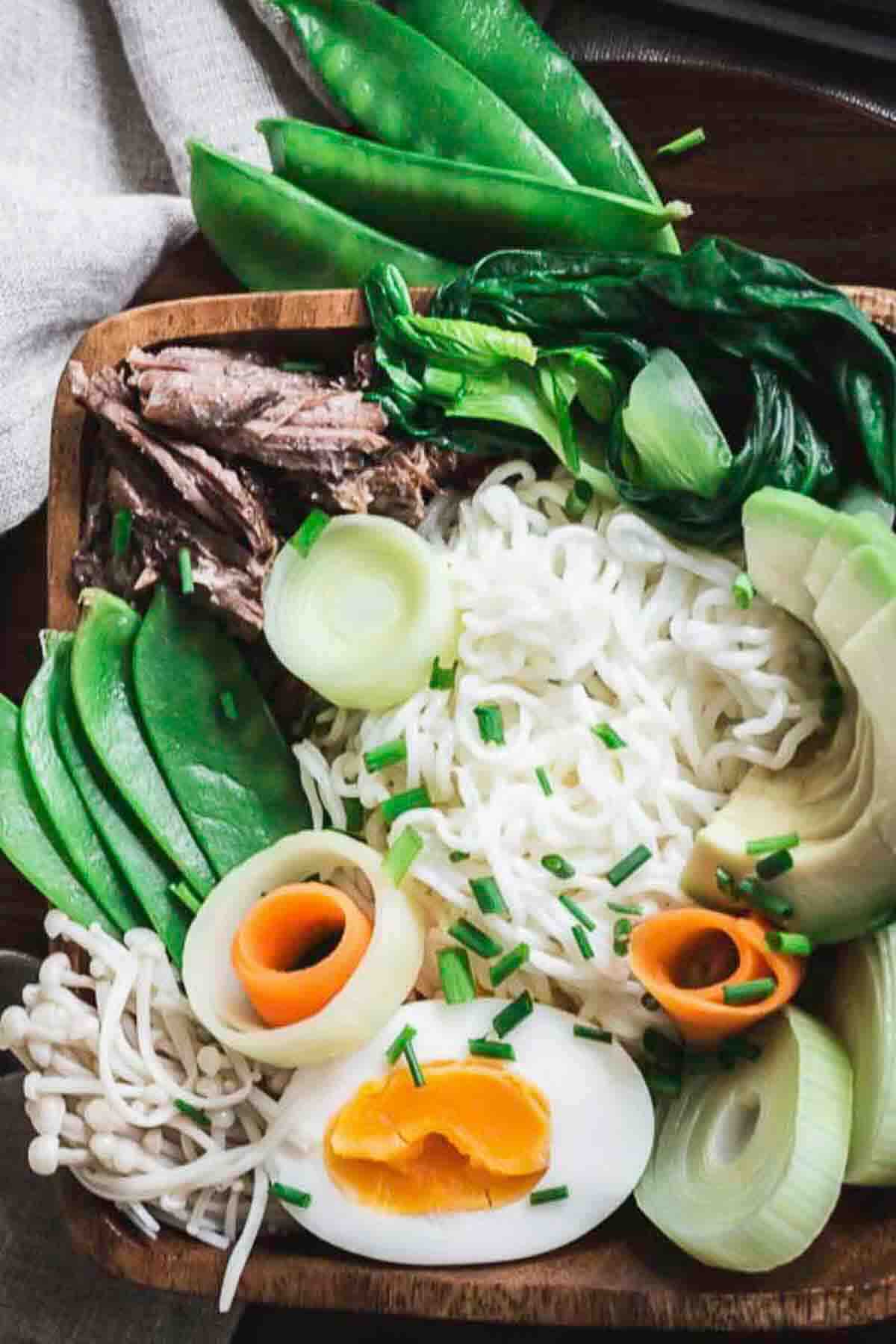 A wooden bowl filled with noodles, boiled egg, avocado, snap peas, bok choy, shredded meat, enoki mushrooms, carrot ribbons, and sliced leeks, garnished with chopped chives.