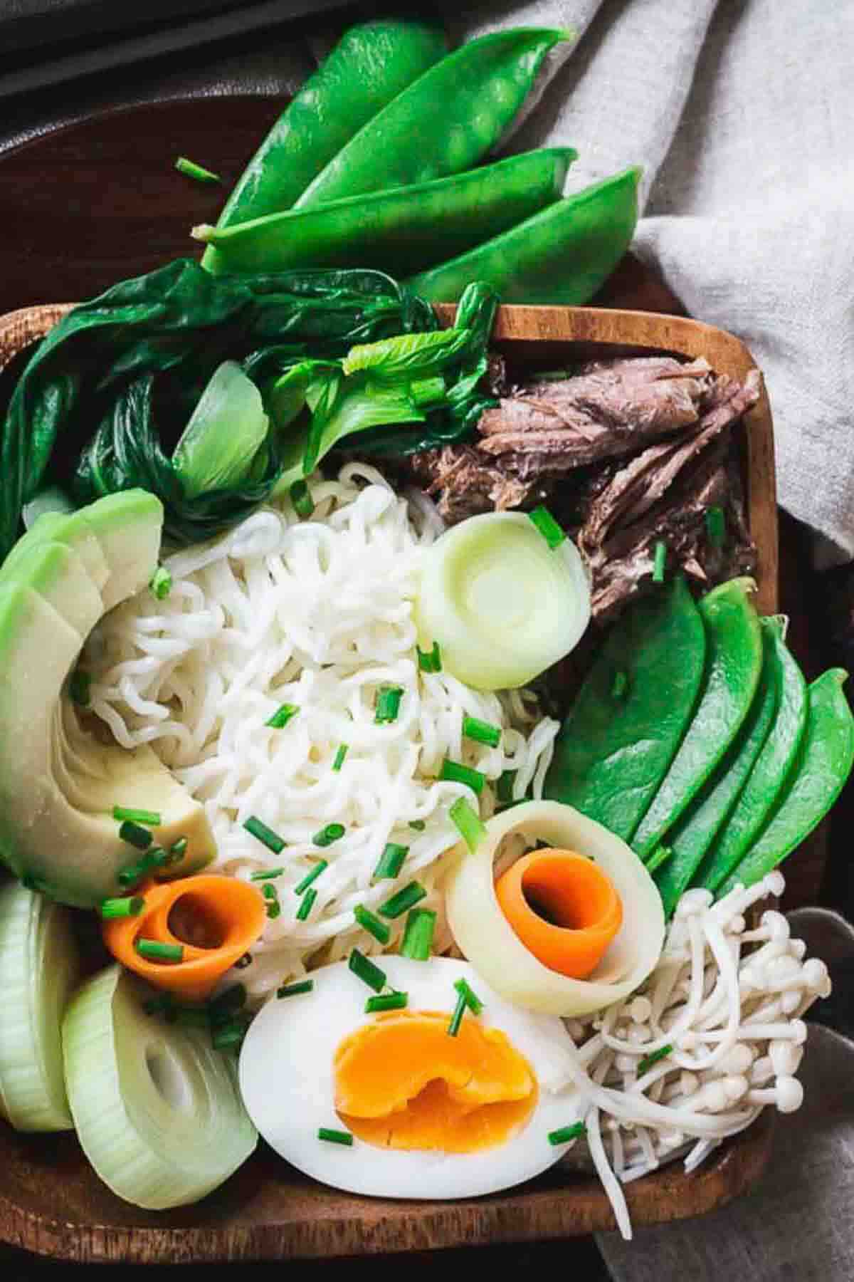 A wooden bowl filled with ramen noodles, sliced avocado, boiled egg, snow peas, leafy greens, shredded beef, enoki mushrooms, and garnished with chopped chives.