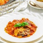 A white plate with a serving of baked eggplant parmesan topped with tomato sauce and melted cheese, garnished with fresh basil leaves. A floral cloth and cutlery are in the background.