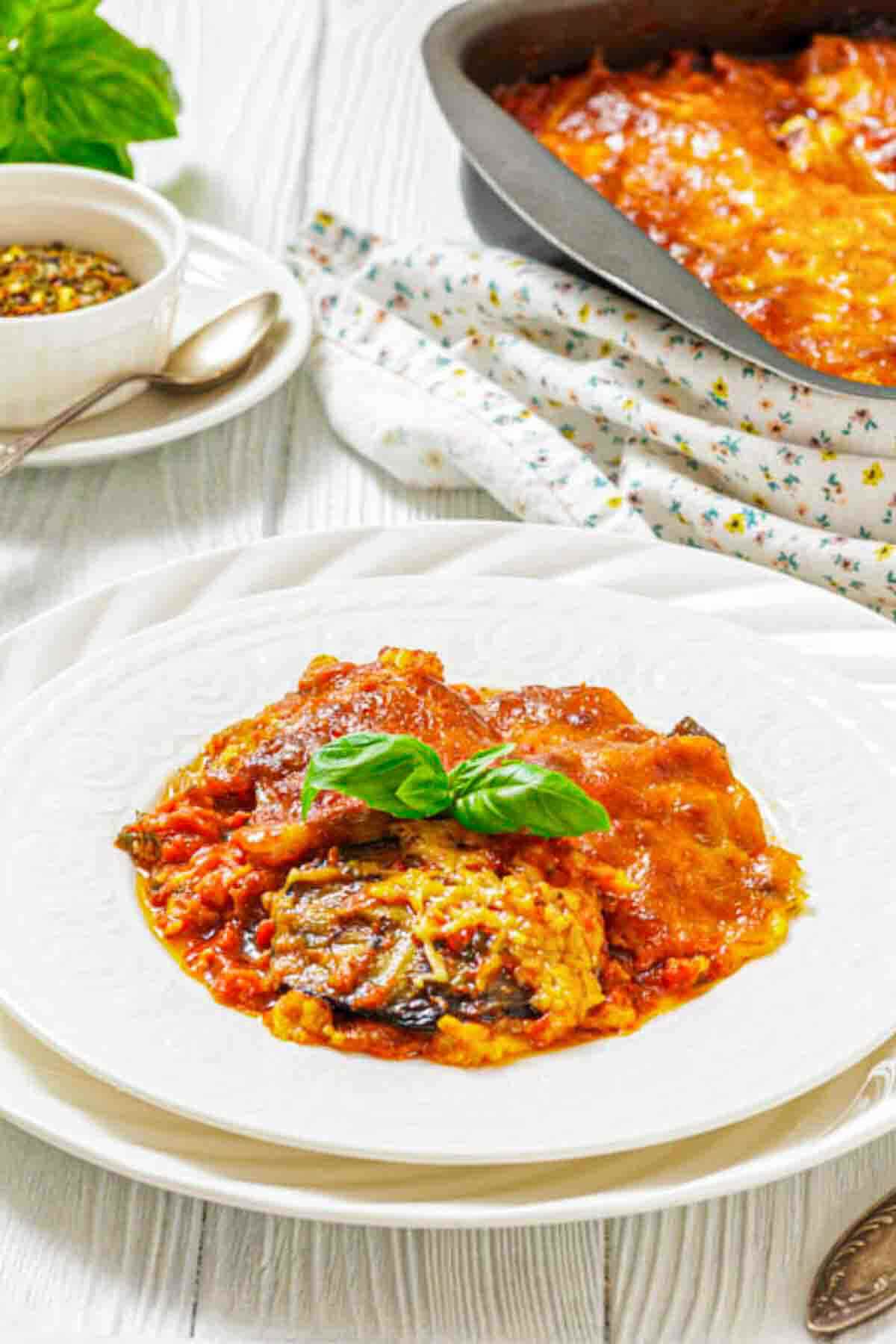 A serving of baked eggplant parmesan topped with basil is plated in the foreground, with a baking dish of the same dish and a bowl of spices in the background.
