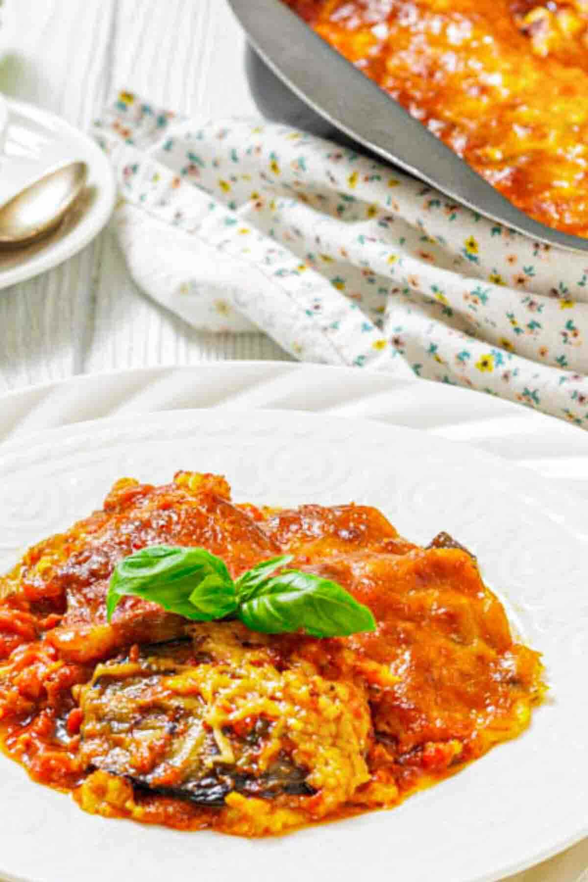 A plate of baked eggplant parmesan with tomato sauce and melted cheese, garnished with fresh basil, sits on a white plate beside a baking dish.