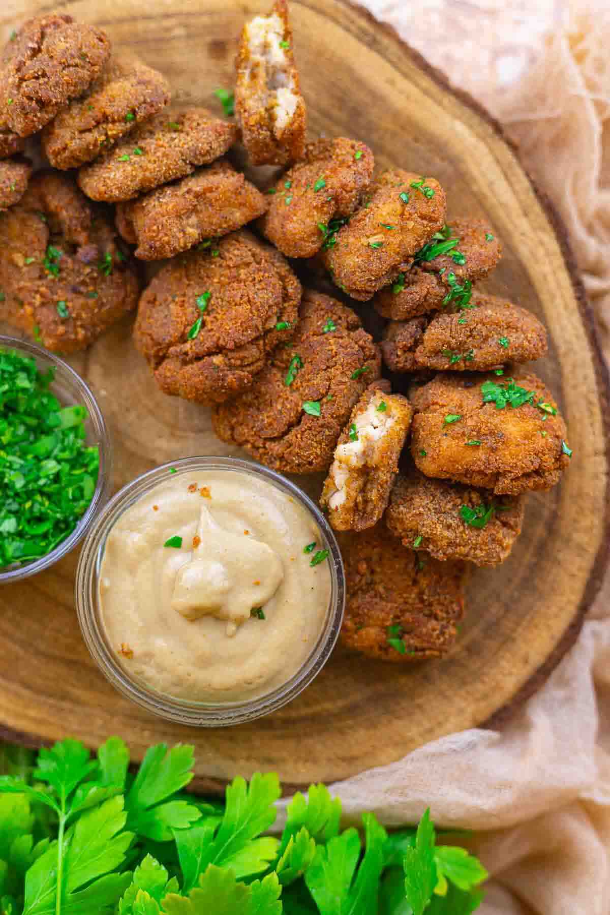 A wooden platter with a row of fried falafel patties, garnished with chopped herbs, served with a bowl of creamy dip and a bowl of fresh chopped parsley.