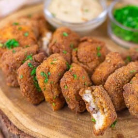 Breaded, fried mushroom bites garnished with chopped herbs, served on a wooden board with dipping sauces in the background.