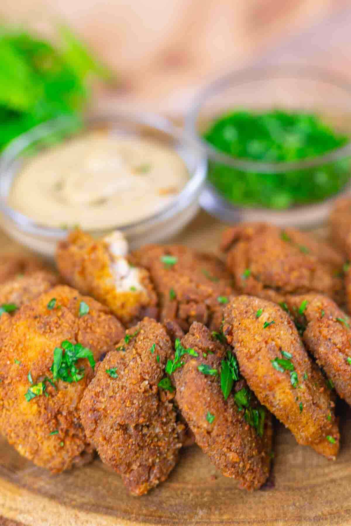 A wooden plate with pieces of breaded, fried chicken garnished with chopped herbs, served with two dipping sauces in small bowls.
