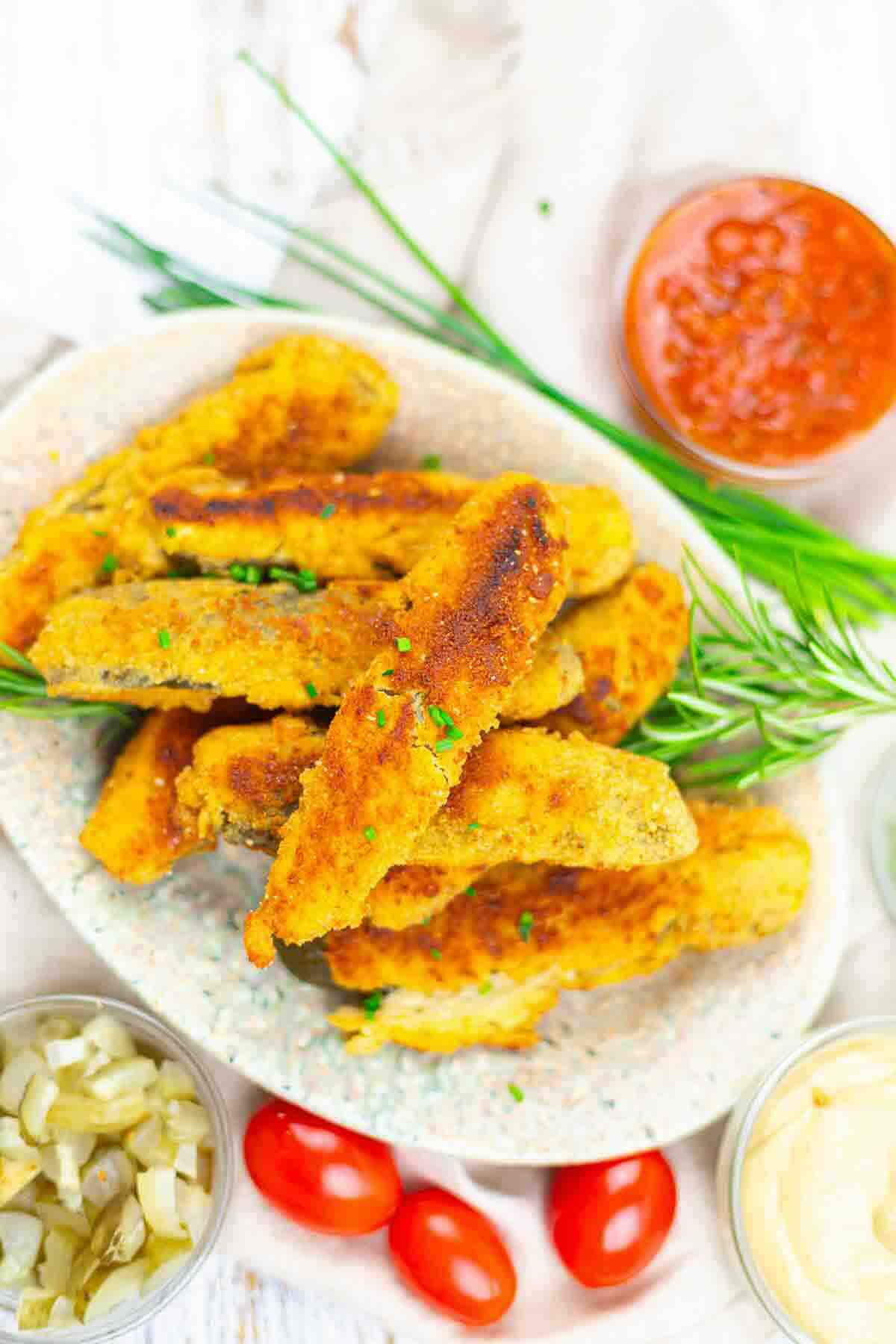 A plate of breaded and fried chicken tenders garnished with herbs, surrounded by bowls of dipping sauces, pickles, and cherry tomatoes.