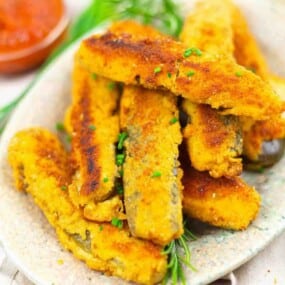 A plate of breaded and fried eggplant sticks garnished with chopped herbs, with a small bowl of red dipping sauce in the background.