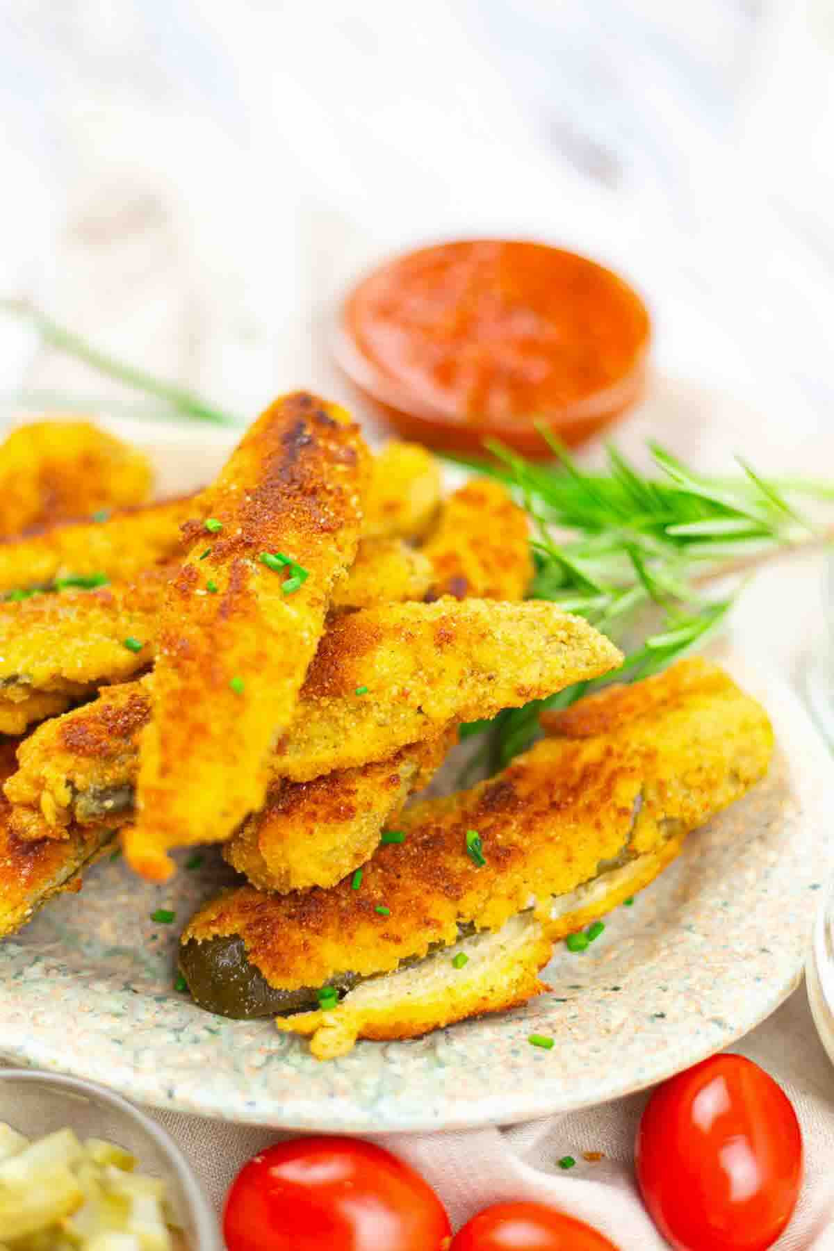 A plate of breaded and fried zucchini sticks garnished with chopped herbs, with a side of red dipping sauce and cherry tomatoes in the background.