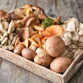 A woven tray filled with a variety of fresh mushrooms, including oyster, cremini, and chanterelle, displayed on a rustic wooden surface.