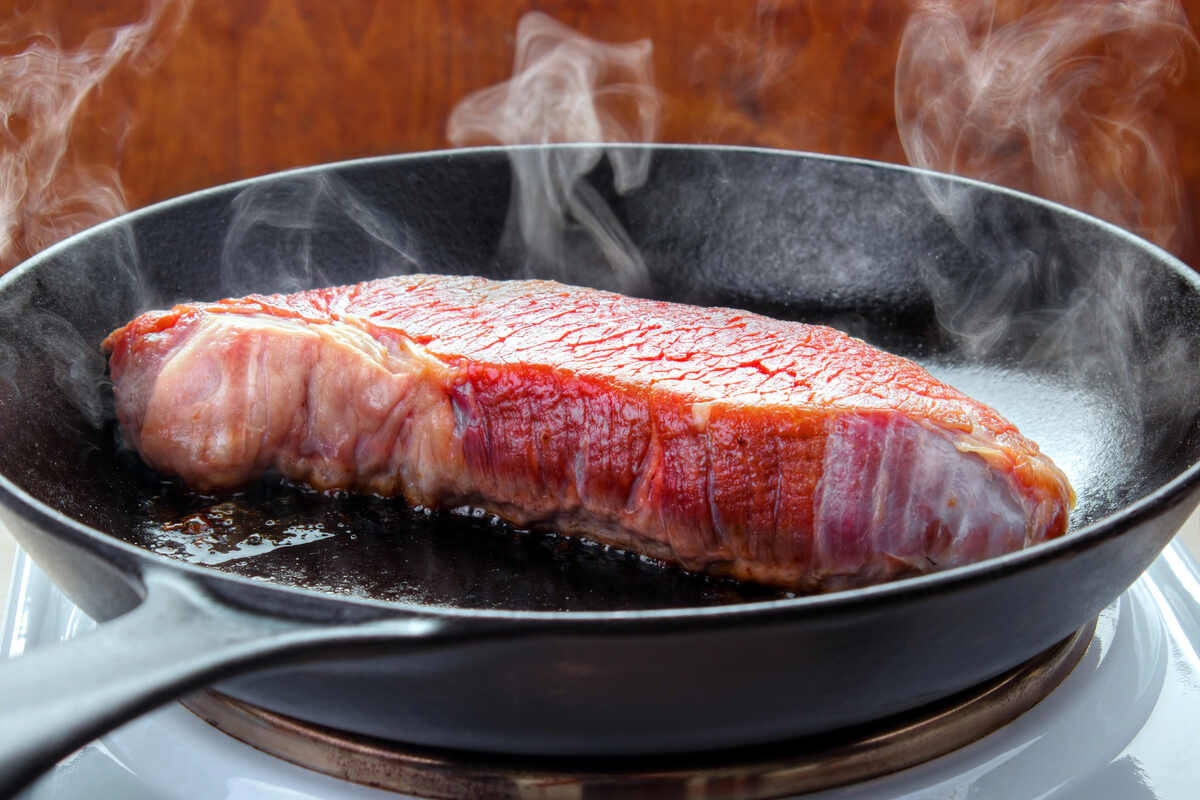 A raw steak is searing in a black frying pan on a stovetop, with steam rising from the meat.