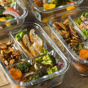Glass meal prep containers filled with grilled chicken, broccoli, carrots, red onion, and pecans arranged in separate compartments on a wooden surface.