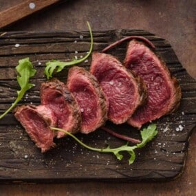 Sliced medium-rare steak arranged on a wooden cutting board with a few arugula leaves and coarse salt sprinkled on top.