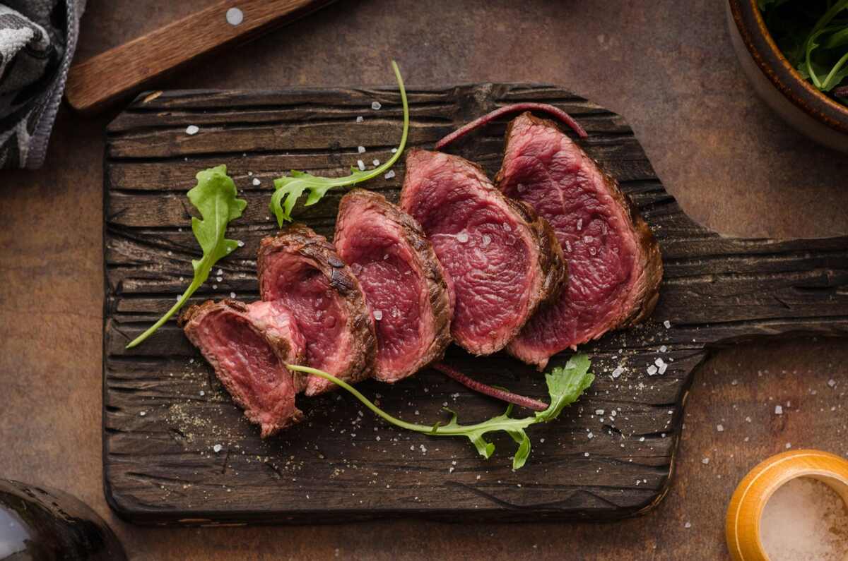 Sliced medium-rare steak arranged on a wooden cutting board with a few arugula leaves and coarse salt sprinkled on top.