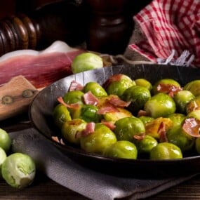 A skillet filled with cooked Brussels sprouts and pieces of ham, surrounded by raw Brussels sprouts, a slice of cured meat, and a red checkered cloth.