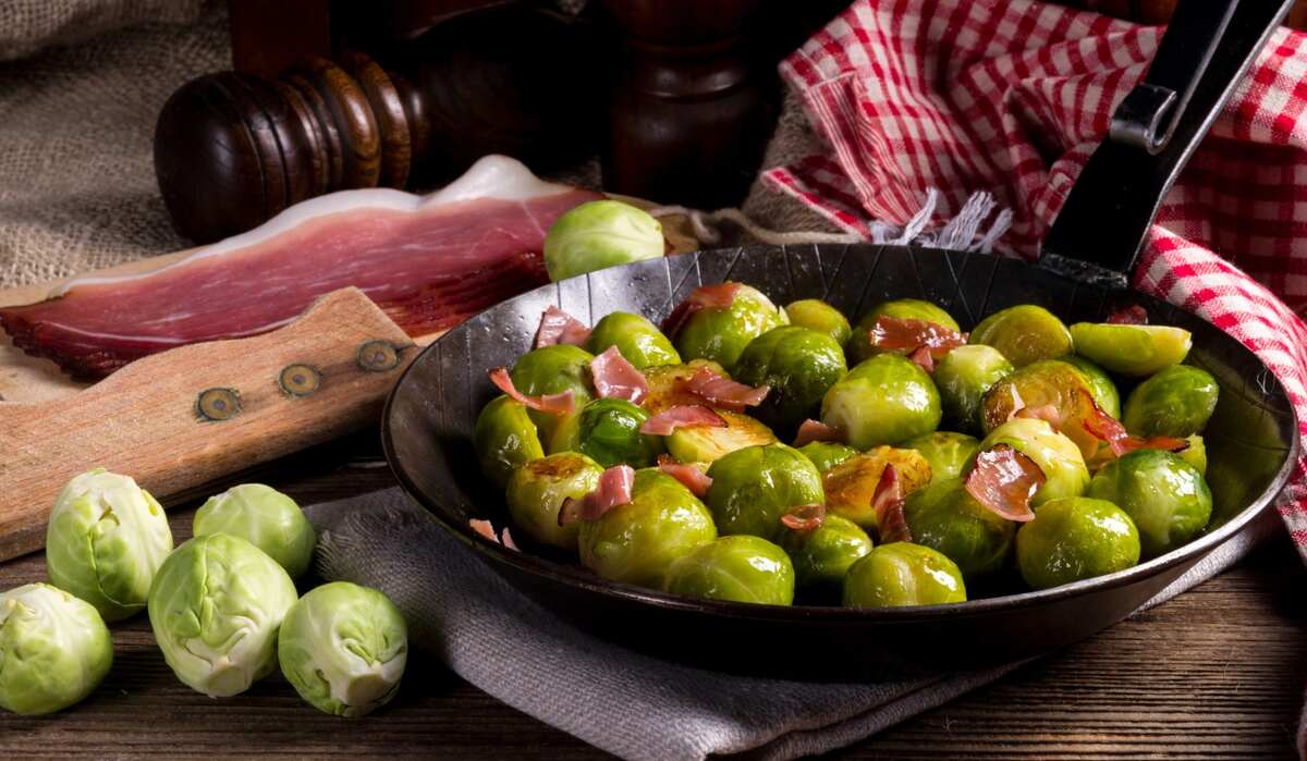 A skillet filled with cooked Brussels sprouts and pieces of ham, surrounded by raw Brussels sprouts, a slice of cured meat, and a red checkered cloth.