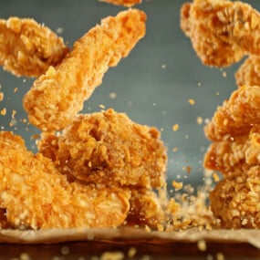 Several pieces of crispy fried chicken are shown mid-air above a brown paper surface, with crumbs scattered around.